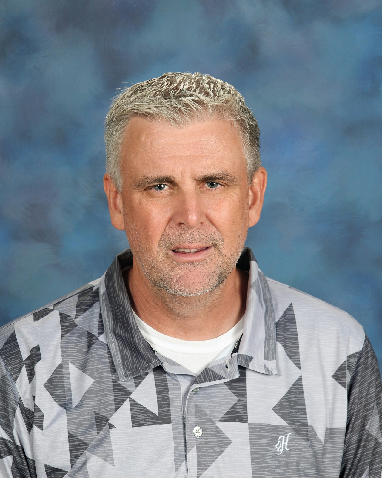 A man with short gray hair is wearing a patterned gray shirt. He has a neutral expression. The background is a smooth blue gradient.